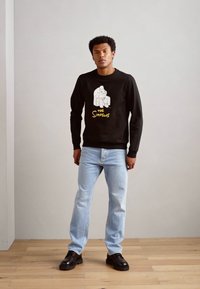 Sudadera negra con un gráfico blanco de Los Simpson, combinada con jeans azul claro y zapatos negros. Suelo de madera y fondo sencillo.