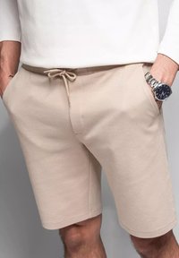 Beige bavlněné šortky s šňůrkou v pase, bočními kapsami a texturovaným povrchem, nošené s bílou košilí s dlouhým rukávem.