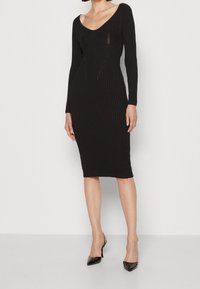 Vestito midi nero a coste con maniche lunghe, scollatura a V profonda e cuciture strutturate. Abbinato a decolté neri a punta con cinturino alla caviglia.