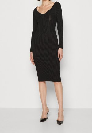 Schwarzes geripptes Midi-Kleid mit langen Ärmeln, tiefem V-Ausschnitt und strukturierten Nähten. Kombiniert mit schwarzen Slingback-Pumps mit spitzem Fuß.