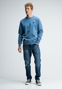 Blauwe katoenen sweatshirt met ronde hals, relaxte pasvorm, geribbelde manchetten en zoom. Gecombineerd met vervaagde blauwe spijkerbroek en zwart-witte gestreepte sneakers.
