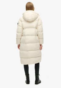 Krämfärgad pufferjacka med huva, med ett quiltat mönster och sidofickor, bärs över svarta leggings och ankelstövlar.