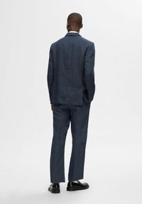 Abito blu navy su misura realizzato in un tessuto strutturato, con uno spacco singolo nella parte posteriore e pantaloni coordinati, indossato con scarpe nere lucide.