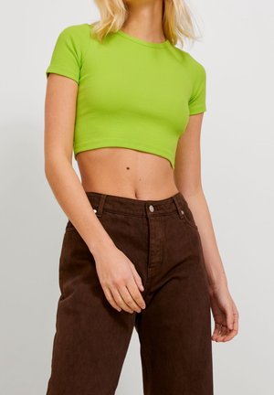 Woman wearing a bright green cropped short-sleeve top and high-waisted dark brown pants, shown from shoulders to hips.