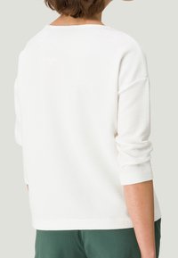 Persona vistiendo una camiseta blanca de manga larga con textura y mangas remangadas, y pantalones verdes, mostrada de espaldas contra un fondo liso.