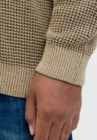 Beez värvi kootud sviiter tekstuuriga mustriga, millel on soonikuga varrukate ääred. Nähtav käsi toetub denimpinnale. Suurendatud vaade toob esile materjali detailid.