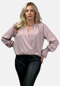 Różowa bluzka z długim rękawem i głębokim dekoltem w kształcie V, z bufkami na rękawach oraz złotym łańcuszkiem przy dekolcie, zestawiona z dopasowanymi czarnymi spodniami.