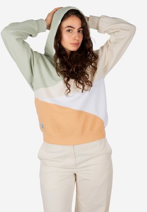 Woman with long curly hair wearing a color-block hoodie in green, beige, white, and orange, with beige pants, posing with hands on her head.
