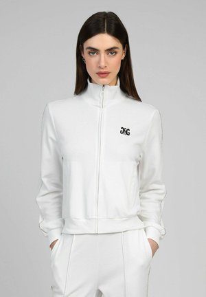 Femme aux cheveux foncés portant une veste blanche à fermeture éclair et un pantalon assorti, debout les mains dans les poches devant un fond gris uni.