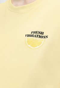 Κίτρινο μπλουζάκι με μαύρο κείμενο "FRESH VIBRATIONS" πάνω από μια εικόνα φέτας λεμονιού στο στήθος, κοντινό πλάνο από τον λαιμό έως τη μέση του κορμού.