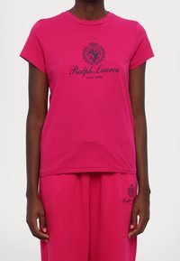 Malli, joka pitää yllään kirkkaan pinkkiä Ralph Lauren -t-paitaa ja siihen sopivia housuja, joissa on laivaston sininen logo ja teksti edessä.