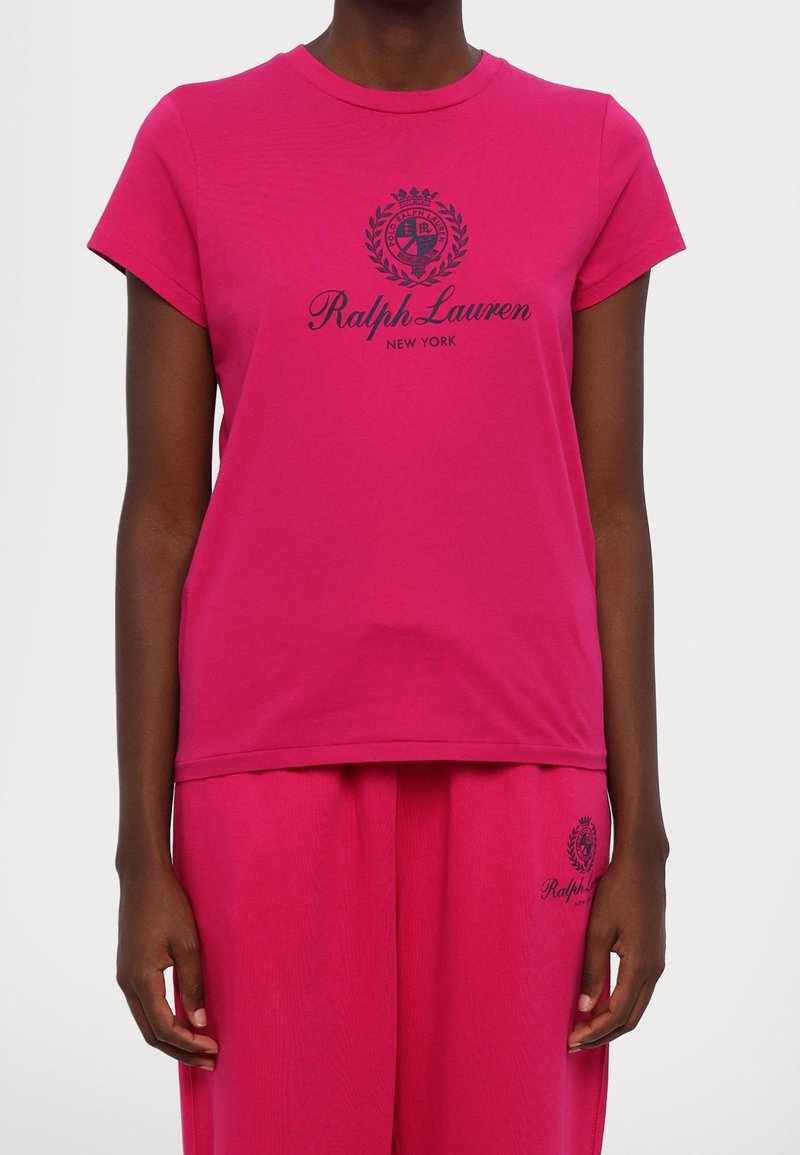 Malli, joka pitää yllään kirkkaan pinkkiä Ralph Lauren -t-paitaa ja siihen sopivia housuja, joissa on laivaston sininen logo ja teksti edessä.