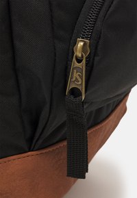 Zaino nero in tessuto con base in pelle marrone, dotato di una cerniera dorata con logo e superficie testurizzata.