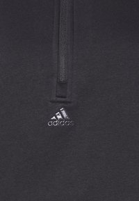 Felpa grigia con zip che presenta un piccolo logo Adidas argentato sul lato sinistro. Si concentra sulla texture del tessuto e sui dettagli della zip.