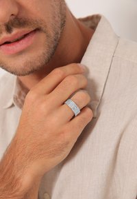 Silberfarbener, strukturierter Ring, der an der rechten Hand getragen wird, mit einem flachen, breiten Band. Der Träger hat hellhäutige Haut und trägt ein Hemd aus Leinen.