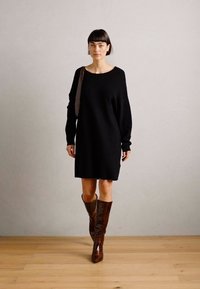 Robe en tricot noir oversize avec épaules tombantes, associée à des bottes en cuir marron arrivant au genou. Texture lisse et coupe décontractée.