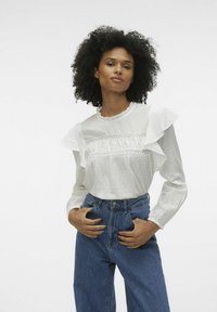 Vit blus med volangärmar och spetsdetaljer, textil med struktur, tillsammans med högmidjade blå denimjeans. Enkel, klassisk design.
