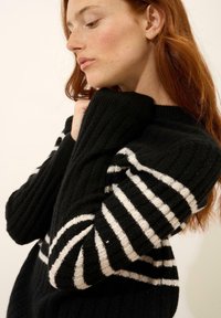 Czarny sweter o sztruksowym splocie z białymi poziomymi paskami. Projekt zawiera wysoki kołnierz i długie rękawy, a także miękką teksturę.