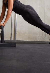 Czarne legginsy sportowe z wzorem teksturowym i markowymi akcentami, zestawione z dopasowaną czarną bluzką, ustawione do pompkowania na macie do ćwiczeń.