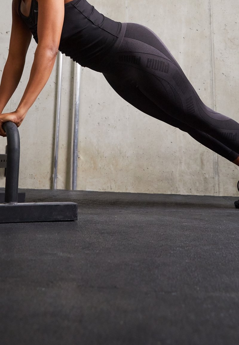 Czarne legginsy sportowe z wzorem teksturowym i markowymi akcentami, zestawione z dopasowaną czarną bluzką, ustawione do pompkowania na macie do ćwiczeń.