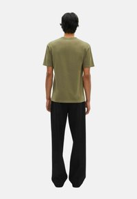 Hombre de pie de espaldas, vistiendo una camiseta de manga corta verde olivo y pantalones negros holgados, frente a un fondo blanco liso.