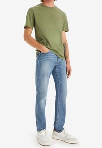 Olivgrön bomulls-t-shirt med en liten röd logga på bröstet, kombinerad med ljusblå denimjeans och vita skor. Avslappnad passform, enkel design.