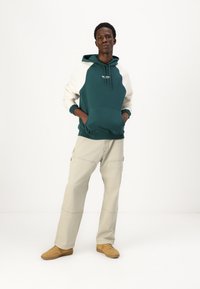 Man standing with hands in pockets wearing teal and white hoodie, beige wide-leg pants, and tan sneakers against white background.