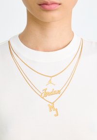 White cotton t-shirt featuring three layered gold necklaces with "Jordan," a jumpman logo, and "MJ" pendants. Smooth texture, casual design.