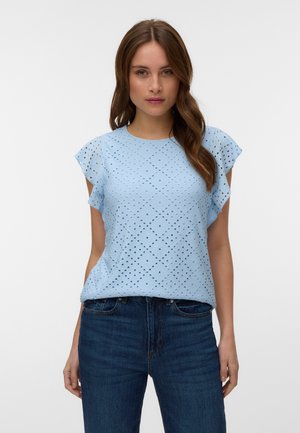 Femme portant un chemisier bleu clair à manches courtes ajourées avec manches à volants et un jean bleu foncé, debout devant un fond clair uni.