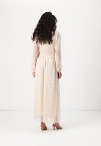 Longue robe beige en tissu transparent à pois, taille élastique et manches longues. Vue de dos montrant une silhouette fluide et une texture douce.