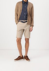 Pantalones cortos beige combinados con una camisa de botones azul oscura y un cárdigan marrón. Zapatillas marrones con suelas blancas. Cinturón trenzado azul.