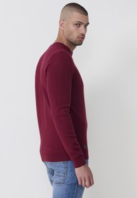 Burgunderfarbener Pullover mit strukturiertem Muster, Rundhalsausschnitt und gerippten Details an den Bündchen und am Saum, kombiniert mit hellblauen Jeans.