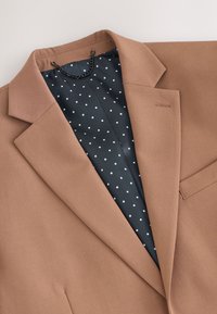 Veste marron avec col, doublure à pois bleu marine. Détails notables : une petite poche discrète et une boucle pour chaîne suspendue.