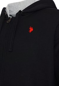 Hanorac negru cu fermoar, căptușit cu un gri pe interiorul glugii, cu șnururi negre și un mic logo brodat roșu al unui jucător de polo pe piept.