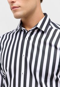 Zwart-wit gestreept shirt met een smal patroon, knoopsluiting met kraag en zilveren knopen. De stof heeft een gladde textuur.