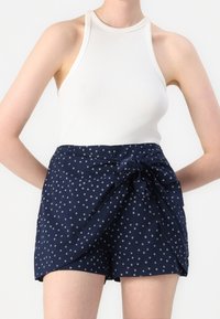 Tmavě modré krátké kalhoty s potiskem, předním zavazováním a bílými kruhovými akcenty. Kombinovány s bílým žebrovaným tílkem bez rukávů. Hladká struktura látky.