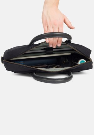 Schwarze Stofftasche mit Ledergriffen, geöffnet, um Innenfächer für Notizbücher, eine Wasserflasche und persönliche Gegenstände zu zeigen.