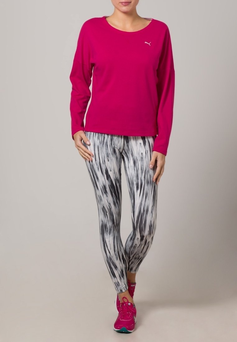 Vaaleanpunainen pitkähiha-paita, jossa on Puma-logo, yhdistettynä musta-valkoisiin kuvioituihin leggingseihin. Mallilla on jalassaan vaaleanpunaiset urheilukengät. Kankaan pinta on sileä.