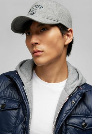 Gray cap with embroidered text, layered over a gray hoodie and navy puffer jacket, worn over a white t-shirt.