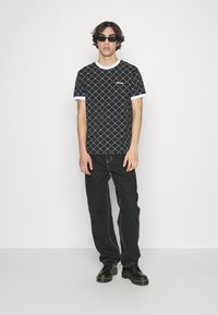Black patterned t-shirt with white accents; geometric design. Worn with black pants and chunky black shoes. Model stands against a neutral backdrop.