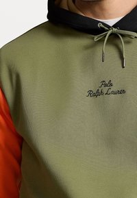 Zbliżenie na osobę wearing oliwkową zieloną i czarną bluzę z kapturem z pomarańczowymi rękawami oraz haftem "Polo Ralph Lauren" na klatce piersiowej.