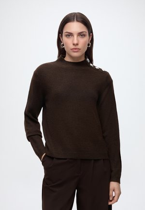 Femme aux longs cheveux foncés porte un pull brun foncé avec des boutons métalliques sur une épaule et un pantalon foncé assorti, debout sur un fond blanc.