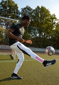 Schwarzes Sportshirt mit drei grünen Streifen, weiße Hose mit schwarzen Seiten und pinken Streifen, schwarze Fußballschuhe, jongliert einen gemusterten Fußball auf Kunstrasen.