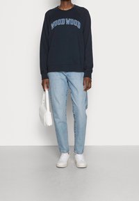 Marineblå sweatshirt med "WOOD WOOD" i vit text, i kombination med ljusblå denimjeans och vita sneakers, hållande en liten vit väska.