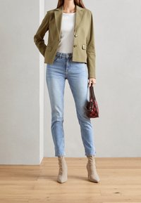 Žena oblečená v jasně olivovém blejzru, bílém topu, světle modrých džínách a béžových kotníkových botách, držící malou červenou kabelku s vzorem, stojící na dřevěné podlaze.