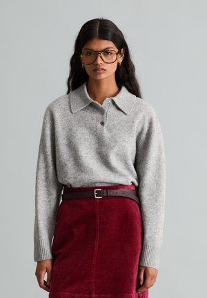 Vrouw met lang donker haar die grote brillen draagt, een grijze trui met kraag en een hoge taille rode ribfluwelen rok met een bruine riem.