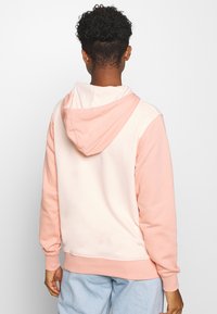 Person med kort, lockigt hår iklädd en tvåfärgad rosa hoodie och ljusblå jeans, stående med ryggen mot betraktaren mot en enfärgad bakgrund.