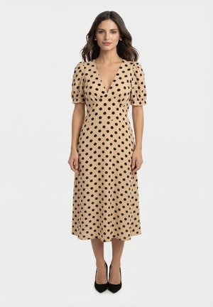 Femme debout portant une robe midi beige à pois noirs, manches courtes bouffantes, décolleté en V, et des talons pointus noirs sur un fond uni.