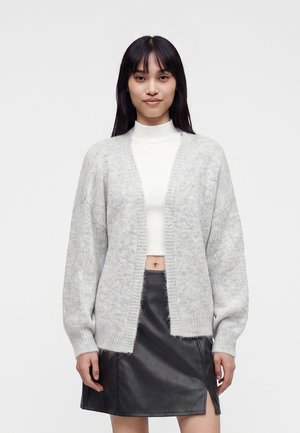 Jeune femme aux cheveux raides et foncés porte un cardigan ouvert gris clair, un col roulé blanc court et une jupe en cuir noire avec une fente à l'avant.