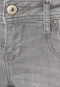 Gros plan d’un jean en denim gris clair montrant une poche avant, une passante de ceinture et un bouton métallique gravé « GENUINE DENIM GOODS ».